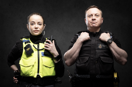 Two officers wearing uniform. One female officer wearing high-visibility vest holding radio. One male officer wearing a black vest and folding arms.