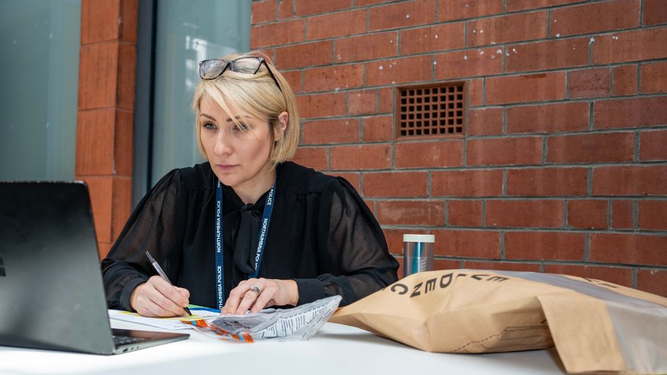 Become a police detective Detective sat at a desk in a police station looking at a laptop surrounded by paperwork and evidence